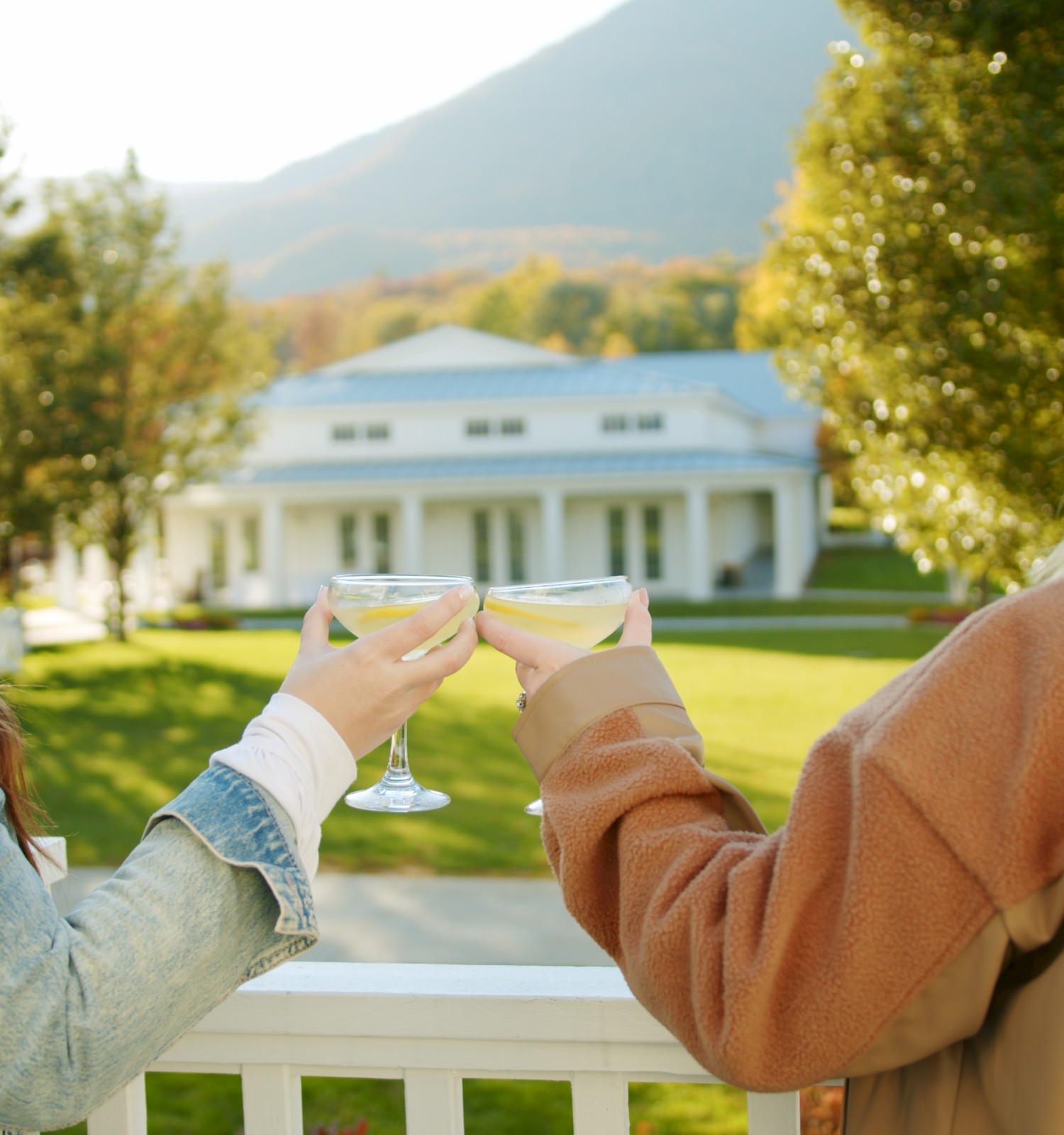 Two people are clinking glasses on a balcony, overlooking a large white house with a scenic background of trees and mountains.
