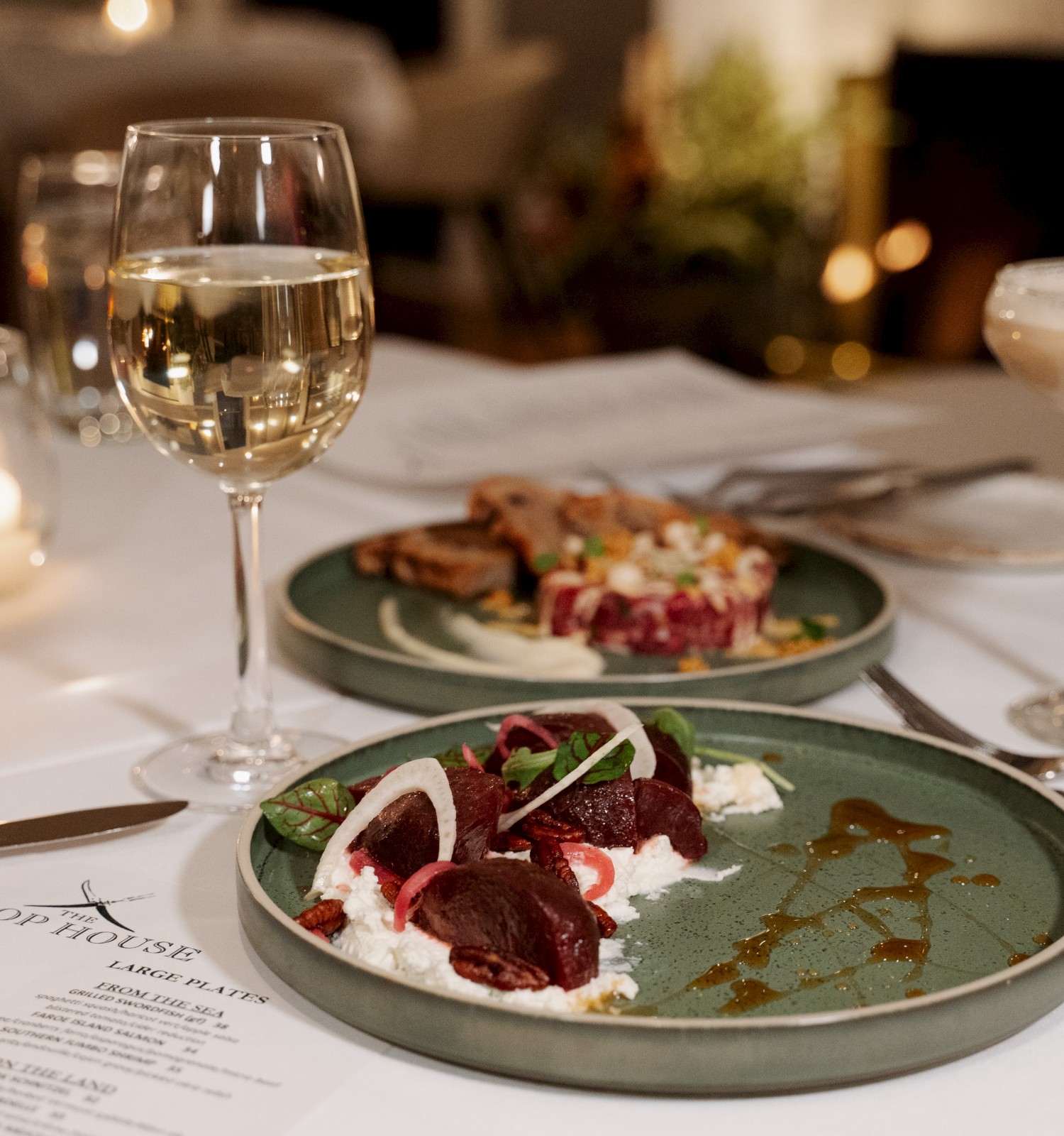 A table set with dishes of food, a glass of white wine, a cocktail, and a menu from The Chop House, lit by a candle.