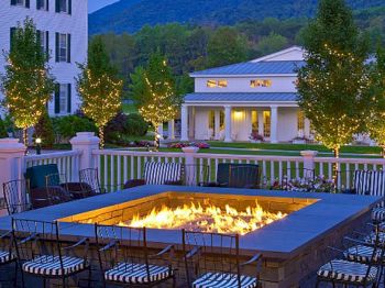 This image features an outdoor patio with a fire pit surrounded by chairs, twinkling lights on trees, and buildings in the background amidst lush greenery.