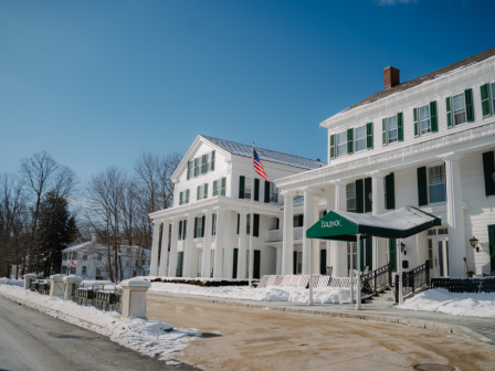 A large, white building with green shutters and a columned entrance, surrounded by snow, displaying an American flag against a blue sky.