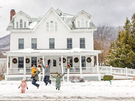 A snowy scene with children playing in front of a large white house decorated for the holidays. Snow is falling and the ground is covered.