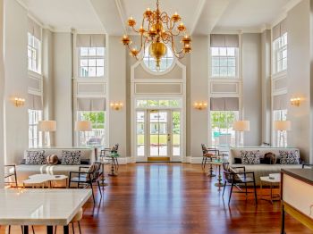 A bright, spacious lobby with high ceilings, large windows, modern furniture, and a central chandelier. Natural light fills the elegant room.