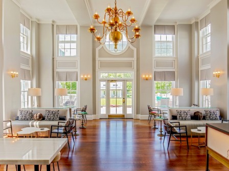 A bright, spacious lobby with high ceilings, large windows, modern furniture, and a central chandelier. Natural light fills the elegant room.