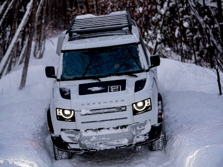 A white SUV driving through a snowy forest path, surrounded by trees and deep snow.