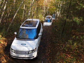 Three white off-road vehicles are driving through a forest trail surrounded by trees and autumn foliage.