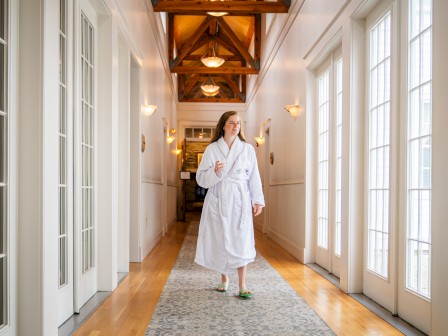 A person in a white robe and slippers walks down a bright hallway with wooden floors and a glass of water in hand.