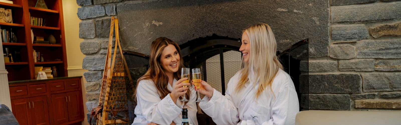Two women in white robes sit on a couch near a cozy stone fireplace, sharing a toast with wine glasses.