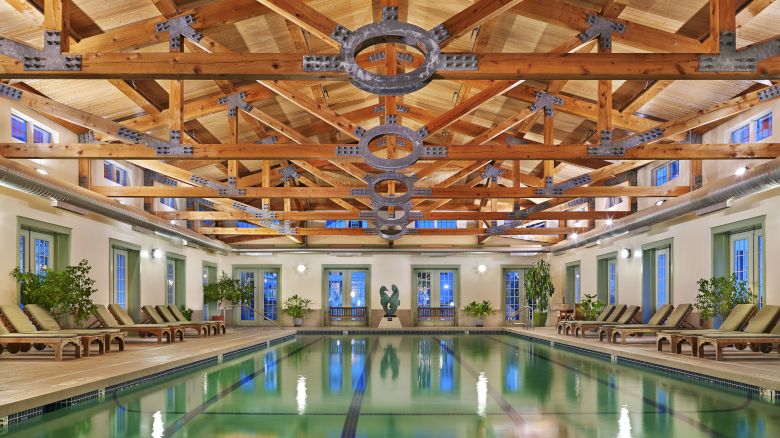 The image shows an indoor pool area with wooden ceiling beams, lounge chairs on the sides, and large windows along the walls, creating a serene atmosphere.