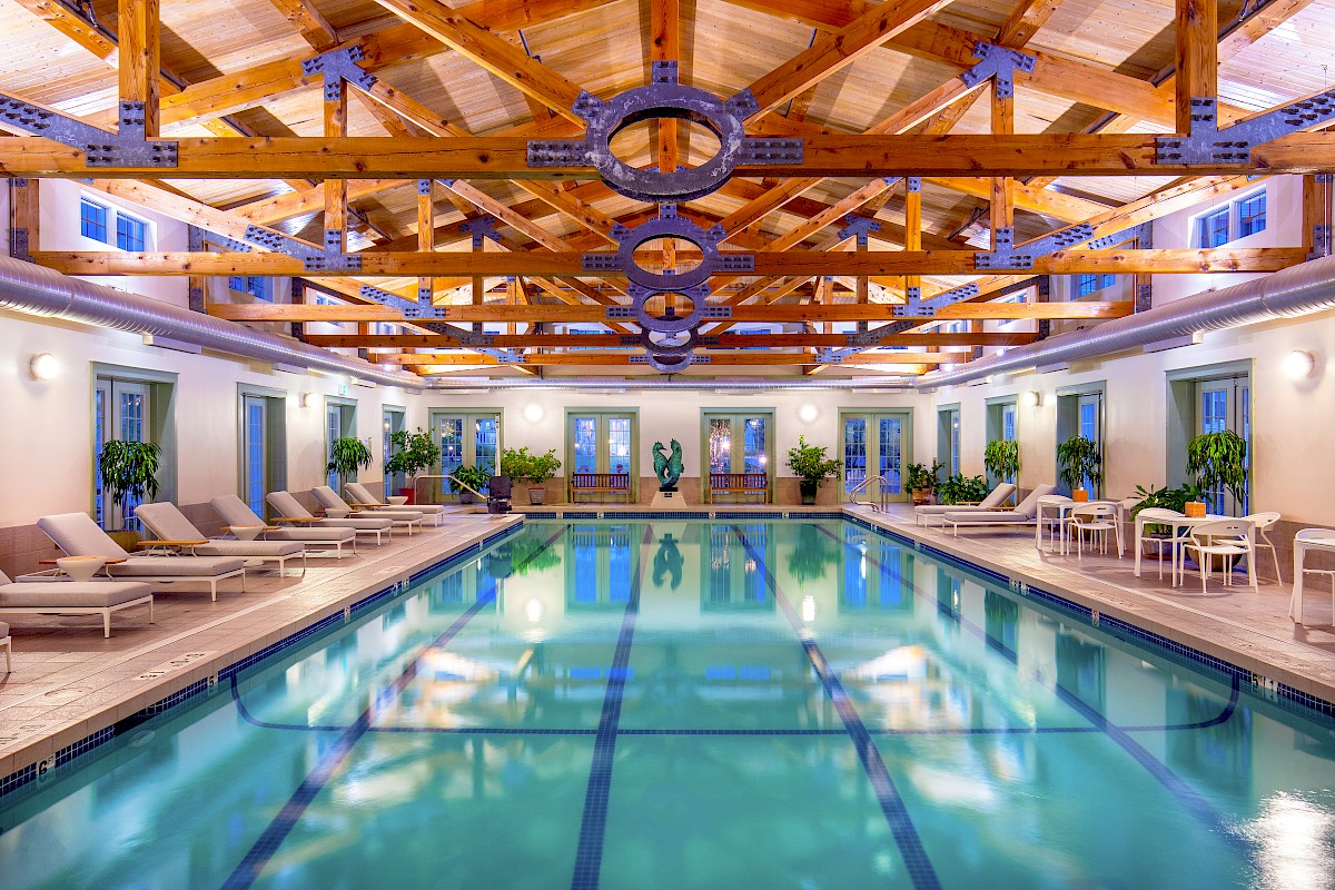 An indoor swimming pool area with lounge chairs, plants, and a wooden vaulted ceiling with metal support beams.