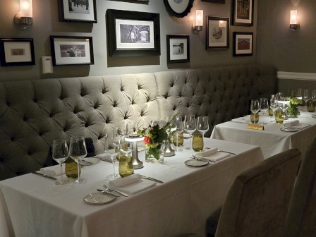 A restaurant setup with white tablecloths, wine glasses, and elegant decor. Framed photos adorn the wall above tufted seating.