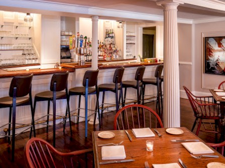 A cozy bar and restaurant with a polished countertop, bottles of alcohol, high chairs, and neatly set tables with plates, cutlery, and candles.