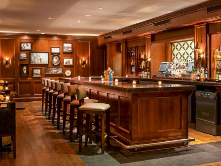 A cozy bar with wooden decor, lined with high chairs, framed pictures on the wall, and shelves stocked with various bottles behind the counter.