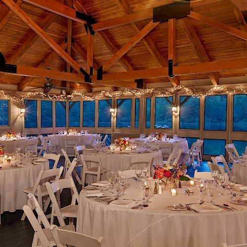 The image shows an elegant reception hall set for an event with round tables, white chairs, candles, and string lights.