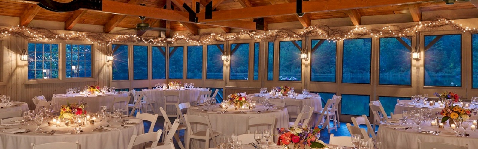 A beautifully decorated dining hall with round tables, white chairs, and warm lighting, set for an event with floral centerpieces and candles.