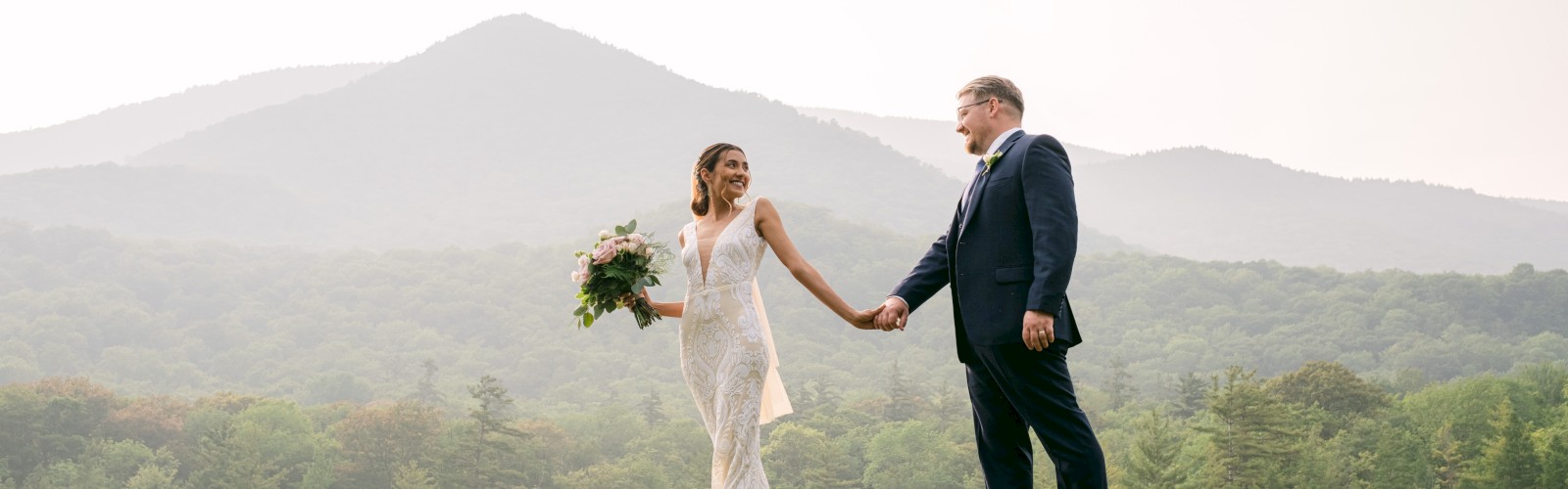 A couple in wedding attire holds hands and walks on grass with a picturesque mountainous landscape in the background.