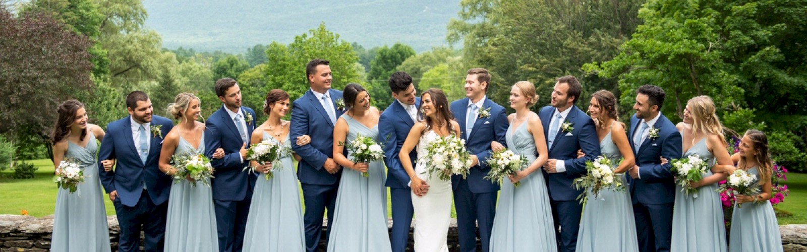 A wedding party stands in a line outdoors; the bride and groom are in the middle, surrounded by bridesmaids in light blue dresses and groomsmen in suits.