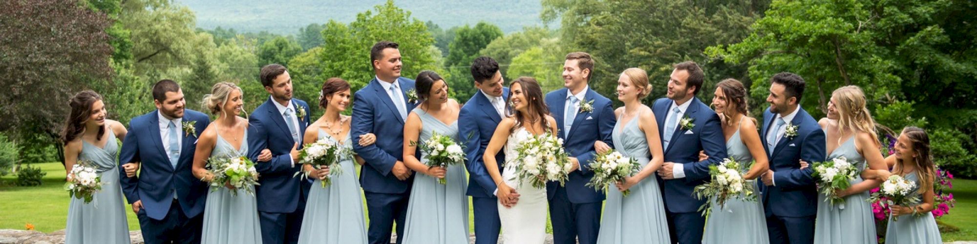 A wedding party stands in a line outdoors; the bride and groom are in the middle, surrounded by bridesmaids in light blue dresses and groomsmen in suits.