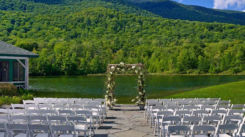 An outdoor wedding setup with rows of white chairs facing a floral arch, set against a backdrop of a lake and green mountains.