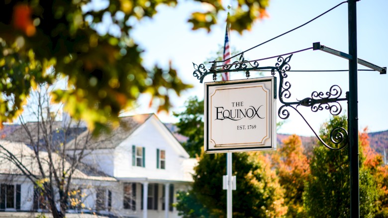 A hanging sign for “The Equinox” on a decorative metal post, with a house and autumn trees in the background, sunny day.