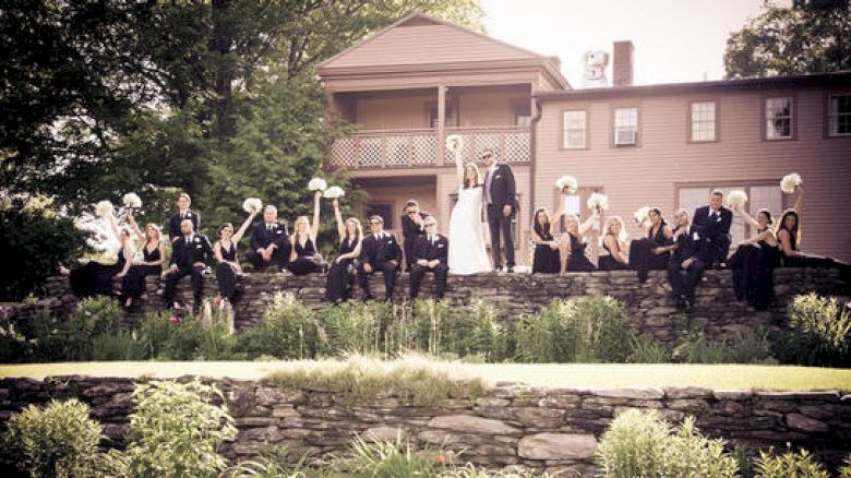 A wedding party poses on a stone wall with greenery, bridesmaids holding bouquets, groomsmen in dark suits, standing by a rustic house.