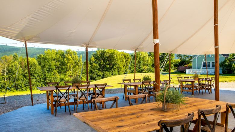 An outdoor seating area under a white tent with wooden tables and chairs, surrounded by greenery and a view of distant hills.
