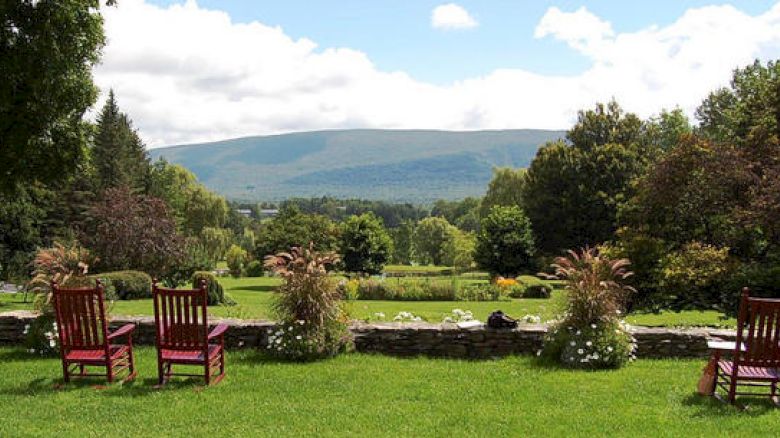 This image features a scenic landscape with wooden chairs on a grassy lawn, overlooking a garden and trees with mountains in the background.