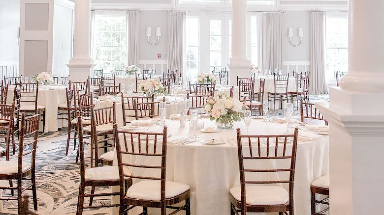 An elegant, light-filled banquet hall with white tablecloths, wooden chairs, floral centerpieces, and a chandelier overhead, ready for an event.
