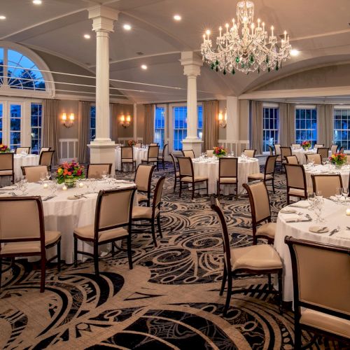 The image shows an elegantly set banquet hall with round tables, floral centerpieces, and chandeliers, prepared for an event or reception.