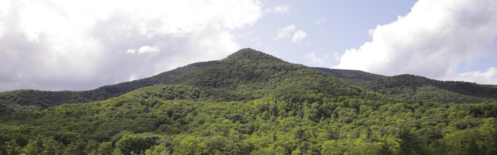The image shows a lush, green mountain range with a clear blue sky and a few scattered clouds. At the base, there's a calm, reflective lake.