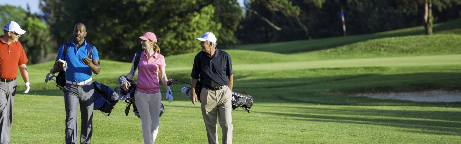 Four people are walking on a golf course, carrying golf bags and wearing casual golf attire, enjoying a sunny day.