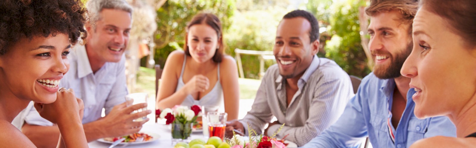 A group of six people are seated around an outdoor table, smiling and enjoying a meal together in a lush green setting, ending the sentence.