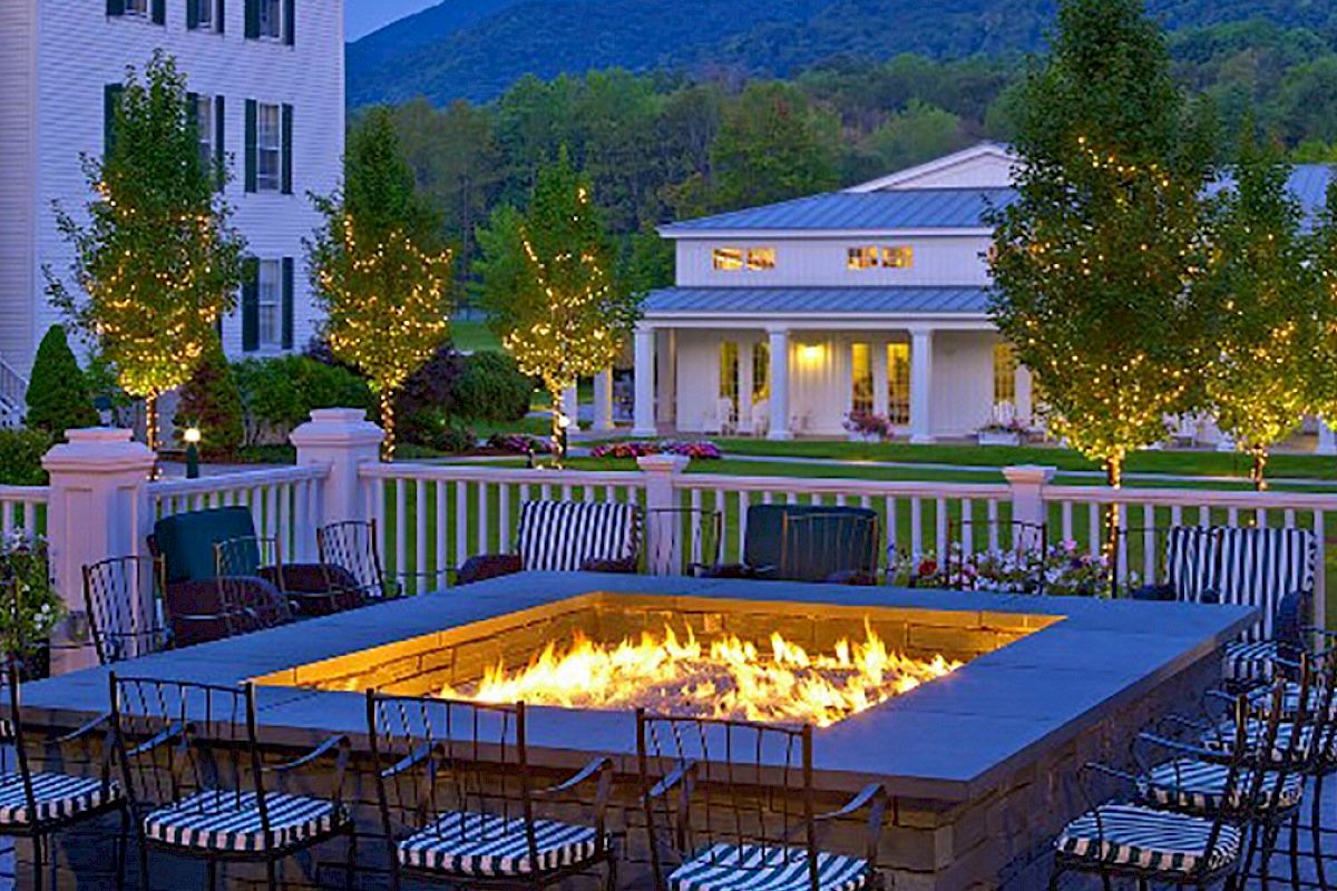 A cozy outdoor area with a fire pit, surrounded by chairs. Fairy lights adorn surrounding trees, and buildings and mountains are in the background.