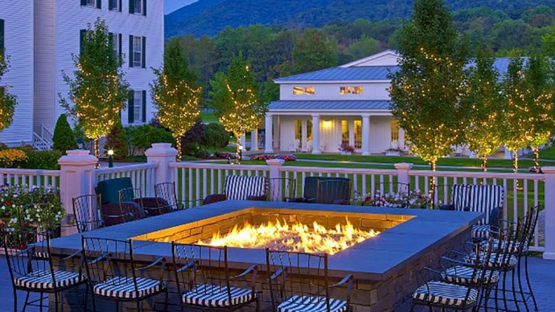 A cozy outdoor area with a fire pit, surrounded by chairs. Fairy lights adorn surrounding trees, and buildings and mountains are in the background.