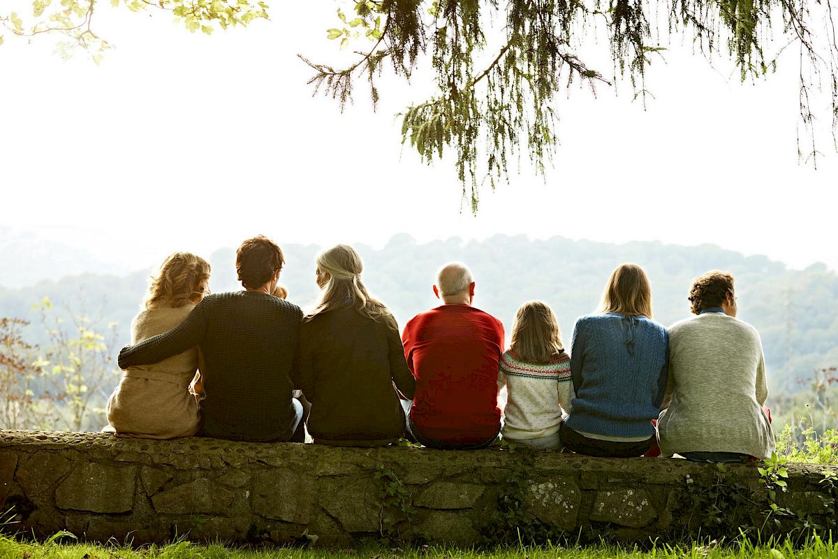 A group of seven people sit on a stone wall, facing away from the camera and looking out at a scenic landscape with trees and rolling hills in the distance.