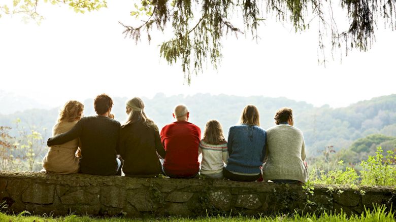 A group of seven people sit on a stone wall, facing away from the camera and looking out at a scenic landscape with trees and rolling hills in the distance.
