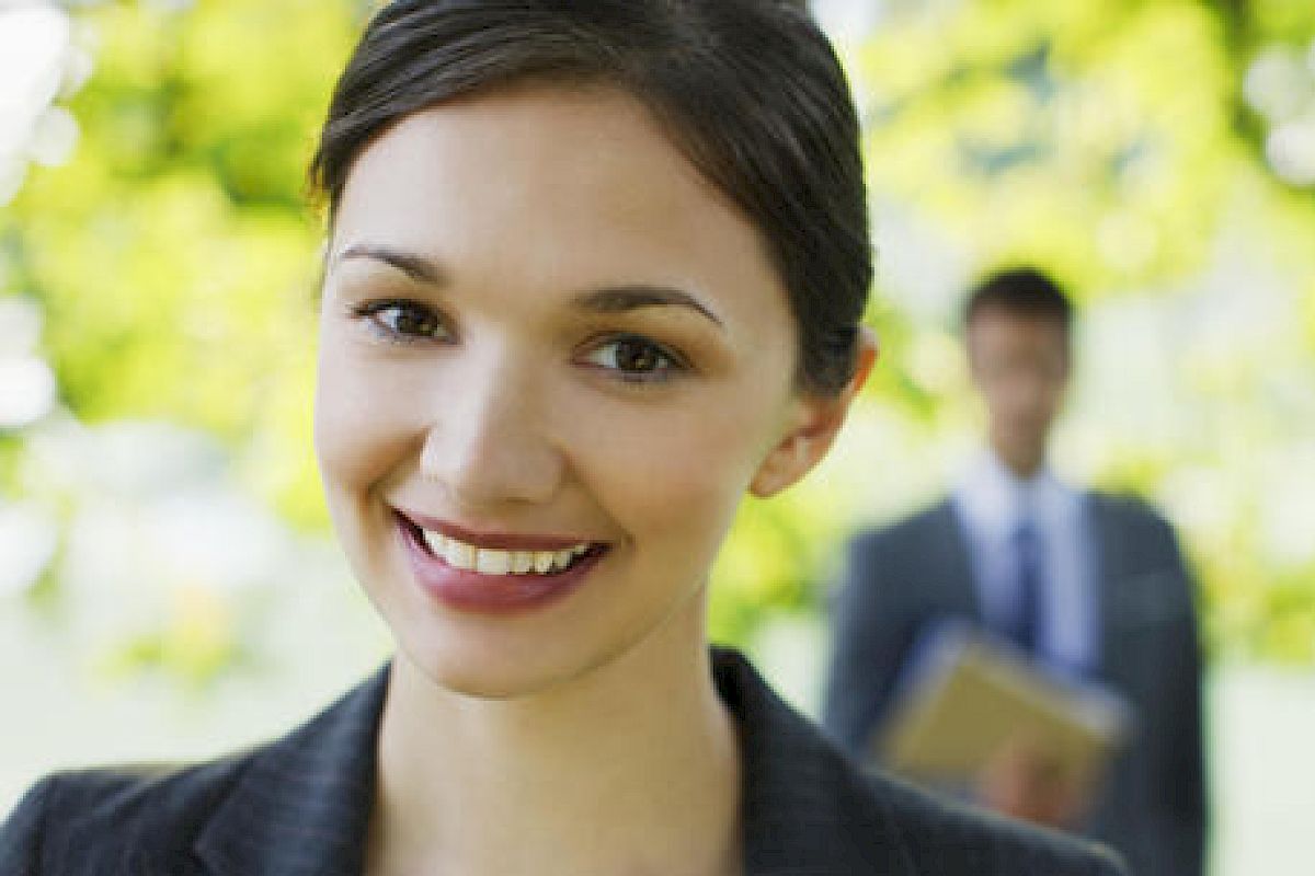 A woman smiling at the camera with a blurred man in a suit standing in the background outdoors, holding some documents.