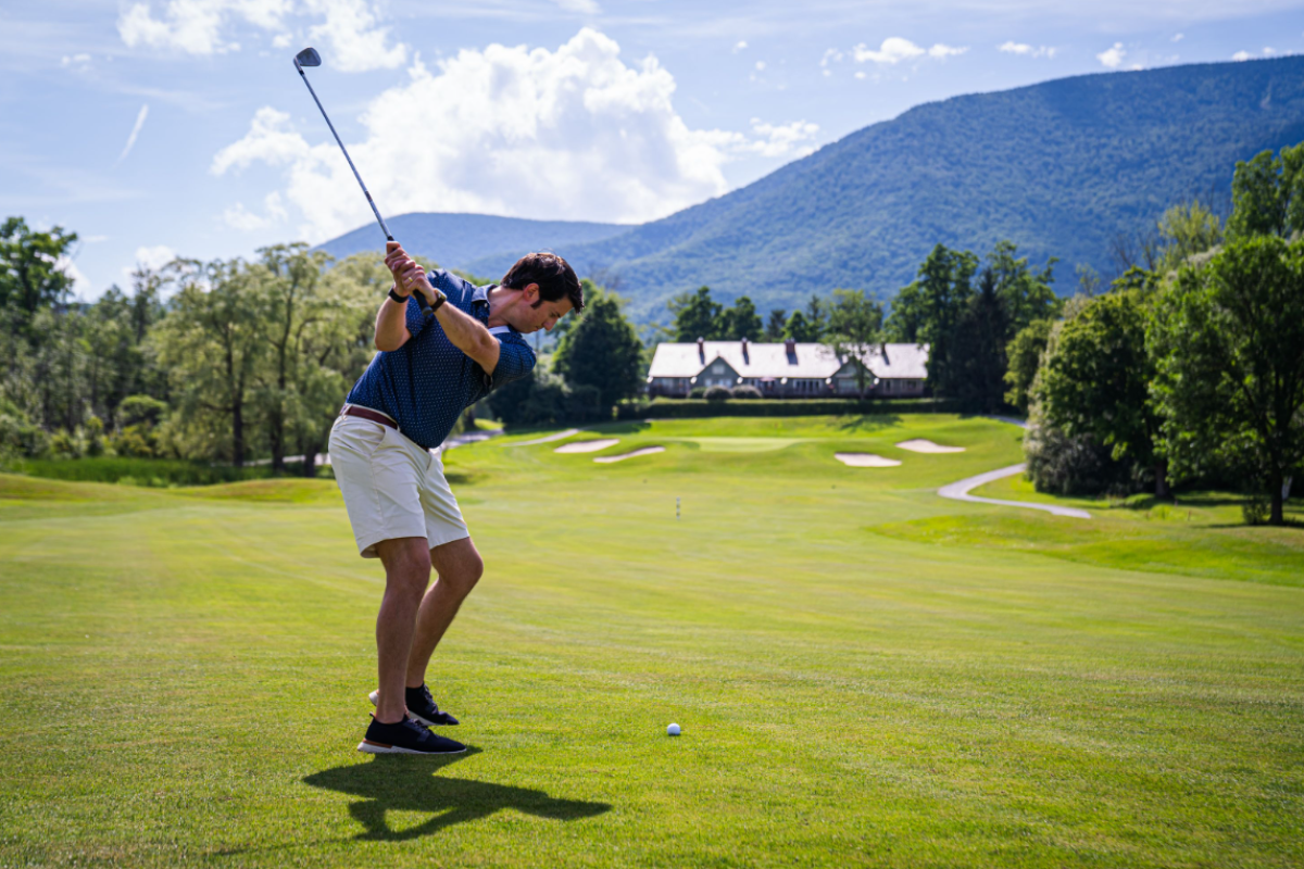 A person is swinging a golf club on a grassy golf course with mountains and a clubhouse in the background.