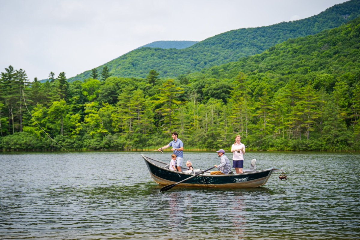 A group of people are fishing from a boat on a lake surrounded by green mountains and dense forest.