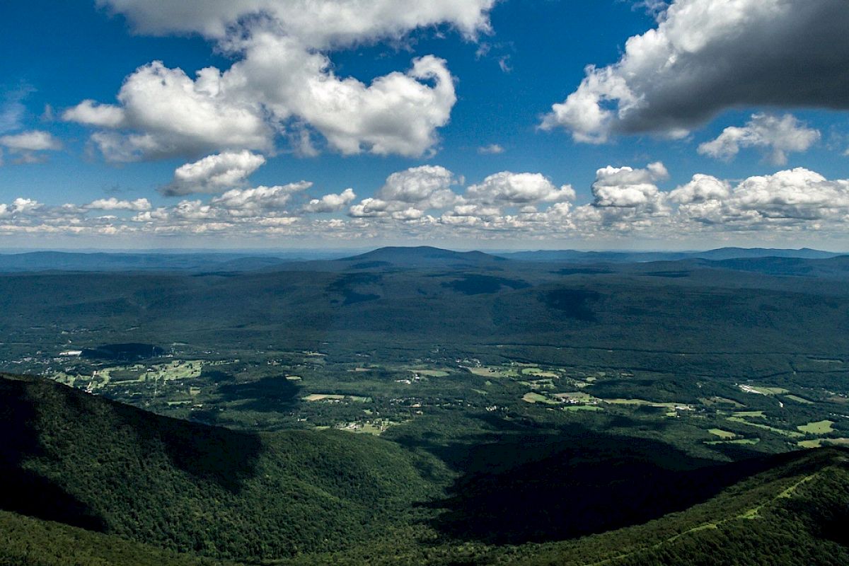 The image shows a vast landscape with rolling green hills, scattered clouds in a blue sky, and a valley with small patches of farmland.