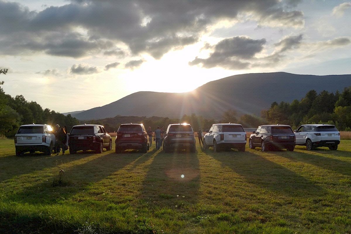 Several SUVs are parked in a row on a grassy field at sunset, with mountains and trees in the background, under a partly cloudy sky.
