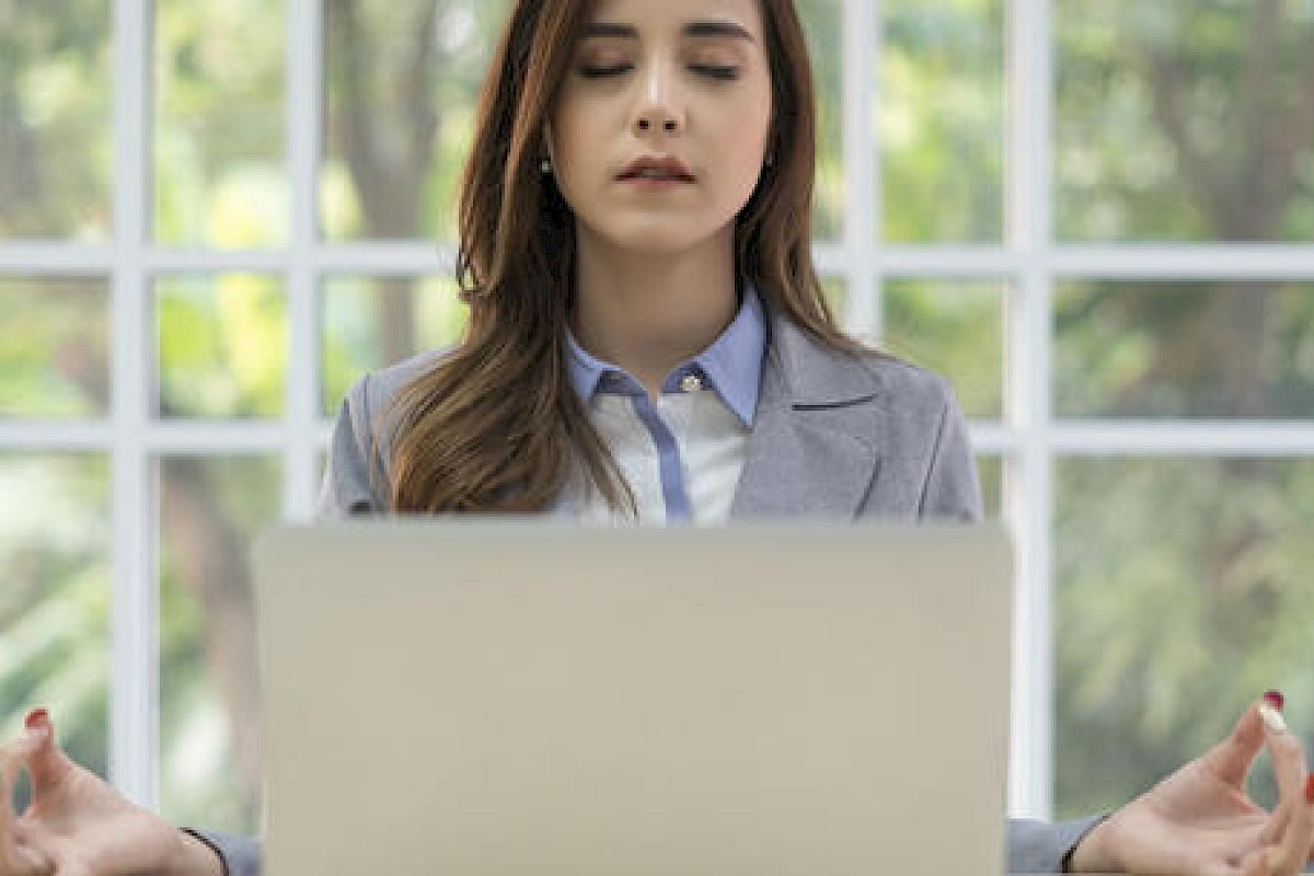A woman sitting with her eyes closed and hands in a meditation pose in front of a laptop, suggesting she is meditating while at work.