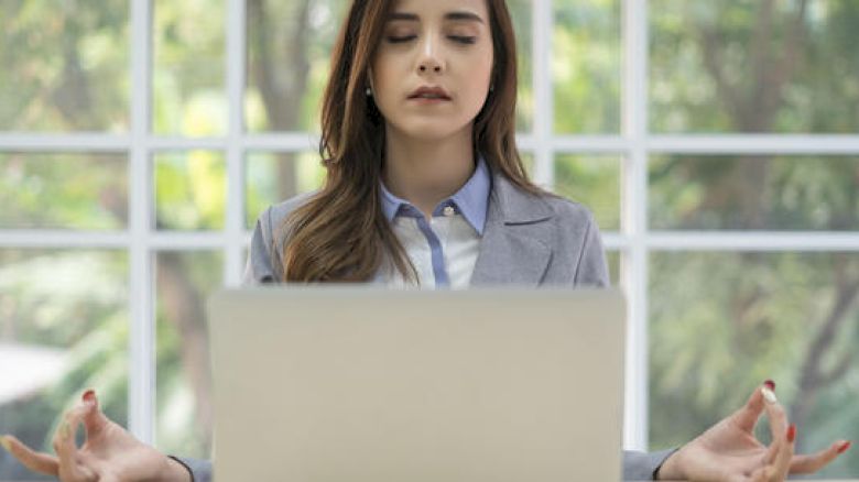 A woman sitting with her eyes closed and hands in a meditation pose in front of a laptop, suggesting she is meditating while at work.
