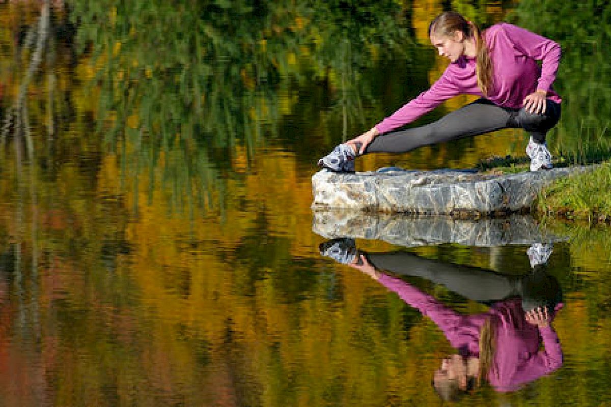 A person is stretching on a rock by a calm lake, with colorful autumn trees reflecting in the water, creating a peaceful and scenic atmosphere.