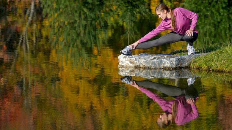 A person is stretching on a rock by a calm lake, with colorful autumn trees reflecting in the water, creating a peaceful and scenic atmosphere.