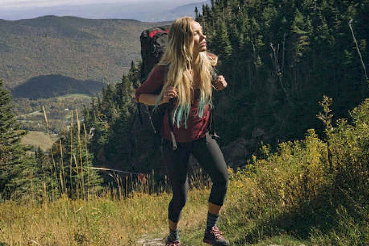 A person with long hair is hiking in a mountainous forested area, carrying a backpack and looking to the side.