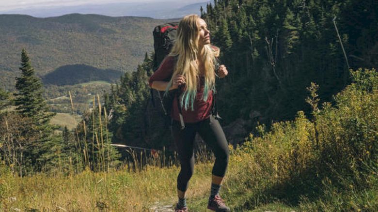 A person with long hair is hiking in a mountainous forested area, carrying a backpack and looking to the side.