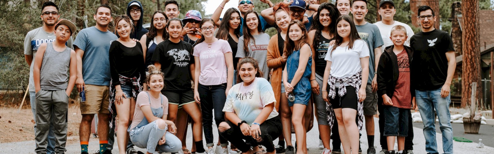 A group of people, including adults and children, are posing for a photo outdoors on a road with trees in the background.