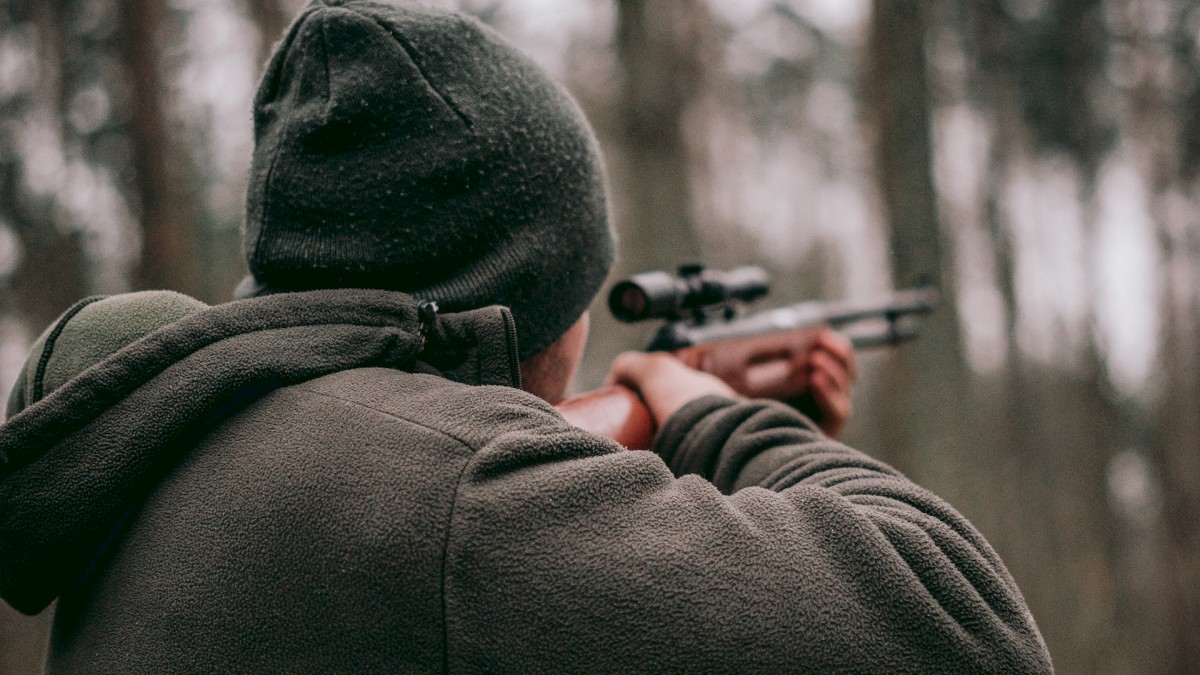 A person wearing a hat and warm clothing is aiming a rifle in a forested area with trees in the background.