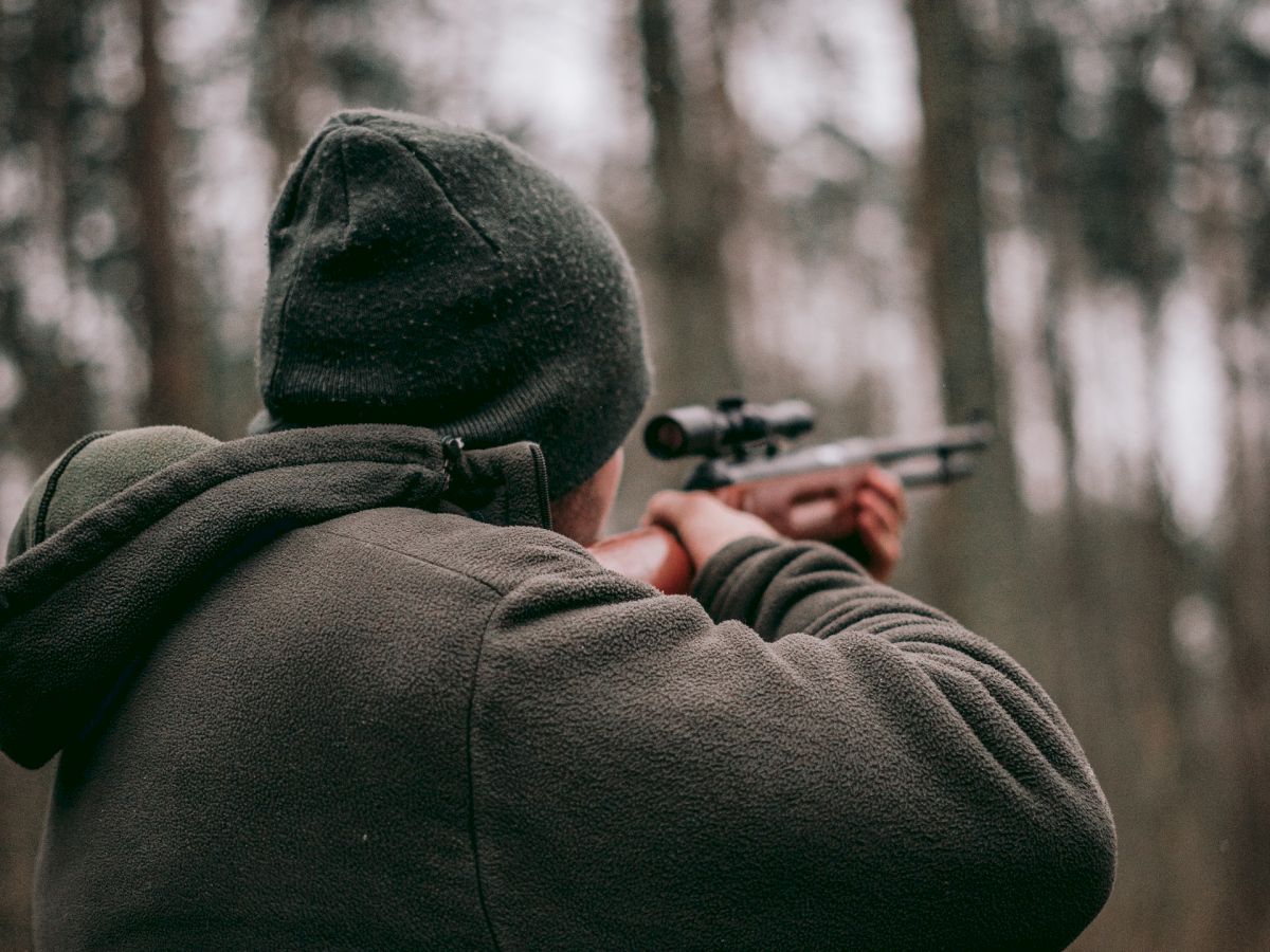 A person wearing a hat and warm clothing is aiming a rifle in a forested area with trees in the background.