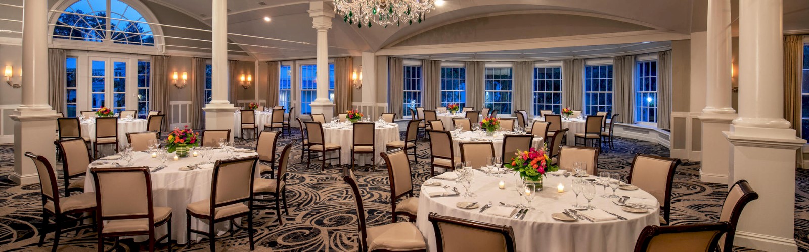 The image shows an elegant banquet hall with round tables set with white linens and floral centerpieces, chandeliers, and decorative wall columns.